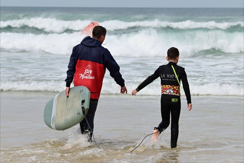 Leçon de surf pour enfant : Moniteur "Surf Moniteur" en action. Moniteur "Surf Moniteur" en veste rouge et élève "LA VIGIE Maison du Surf" en combinaison léopard entrent dans l'océan avec une planche.