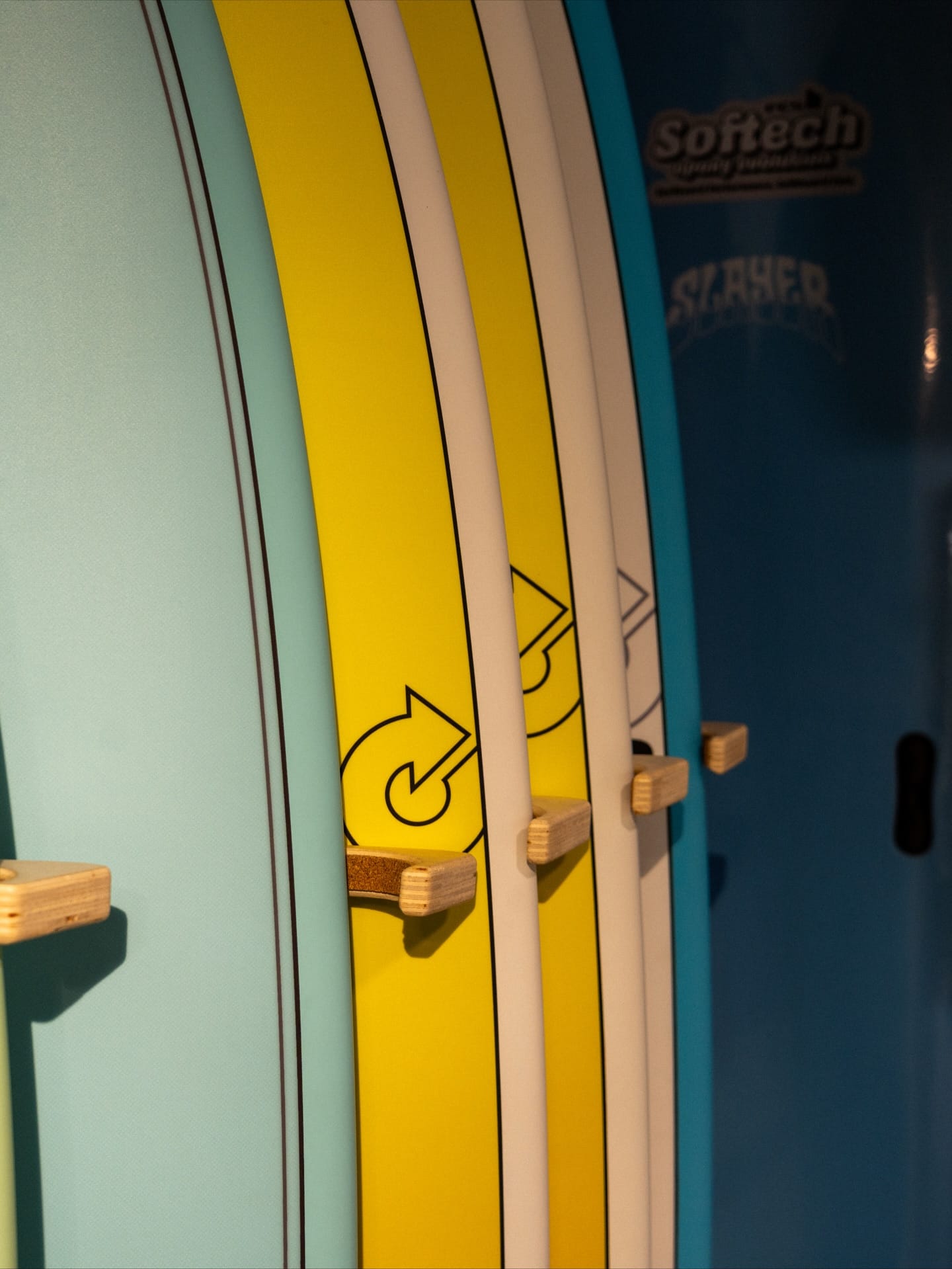 Cinq planches de surf colorées (bleu, jaune, blanc) sur des racks en bois. Logos Softech et Slayer visibles sur le panneau bleu marine.