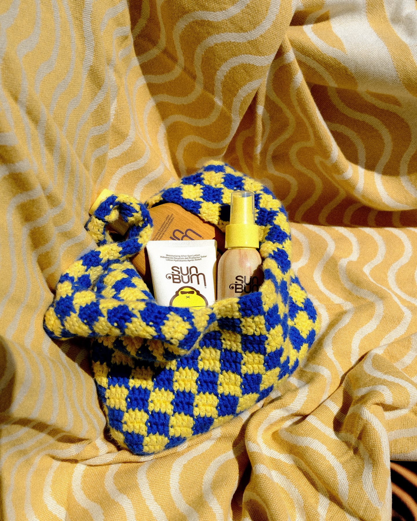 Sac au crochet jaune et bleu à damier contenant une lotion Sun Bum Hydratante Après-Soleil sur un tissu jaune à vagues.