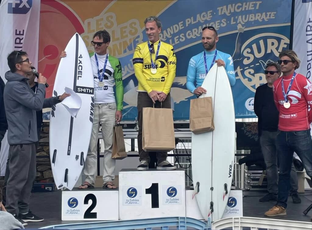 Surf Sables d'Olonne : Les vainqueurs sur le podium. Remise des prix sur le podium de surf Les Sables d'Olonne. Trois surfeurs médaillés avec planches (Excel, Vince) et prix.