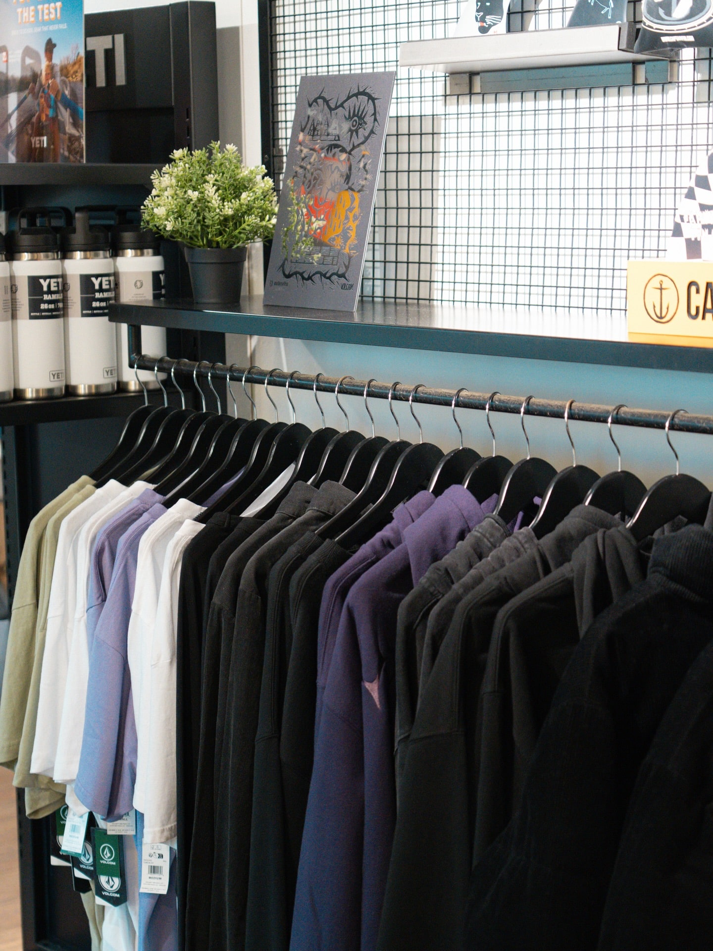 Présentoir de vêtements Volcom colorés sur cintres, du blanc au noir, dans un magasin. Étagère supérieure : bouteilles YETI Rambler, plante et affiche.