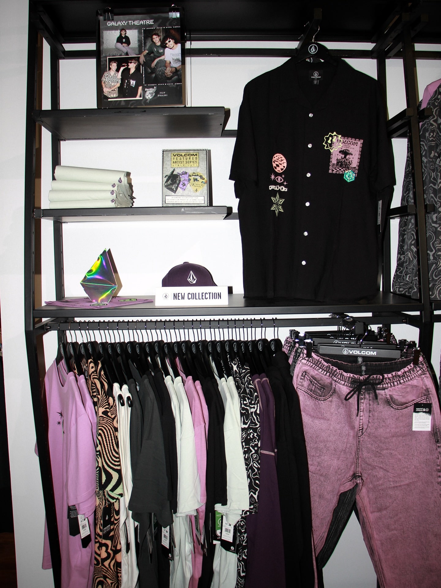Volcom New Collection: chemise noire à patchs, casquette violette, joggers rose délavé. Racks de t-shirts streetwear et posters artistiques.