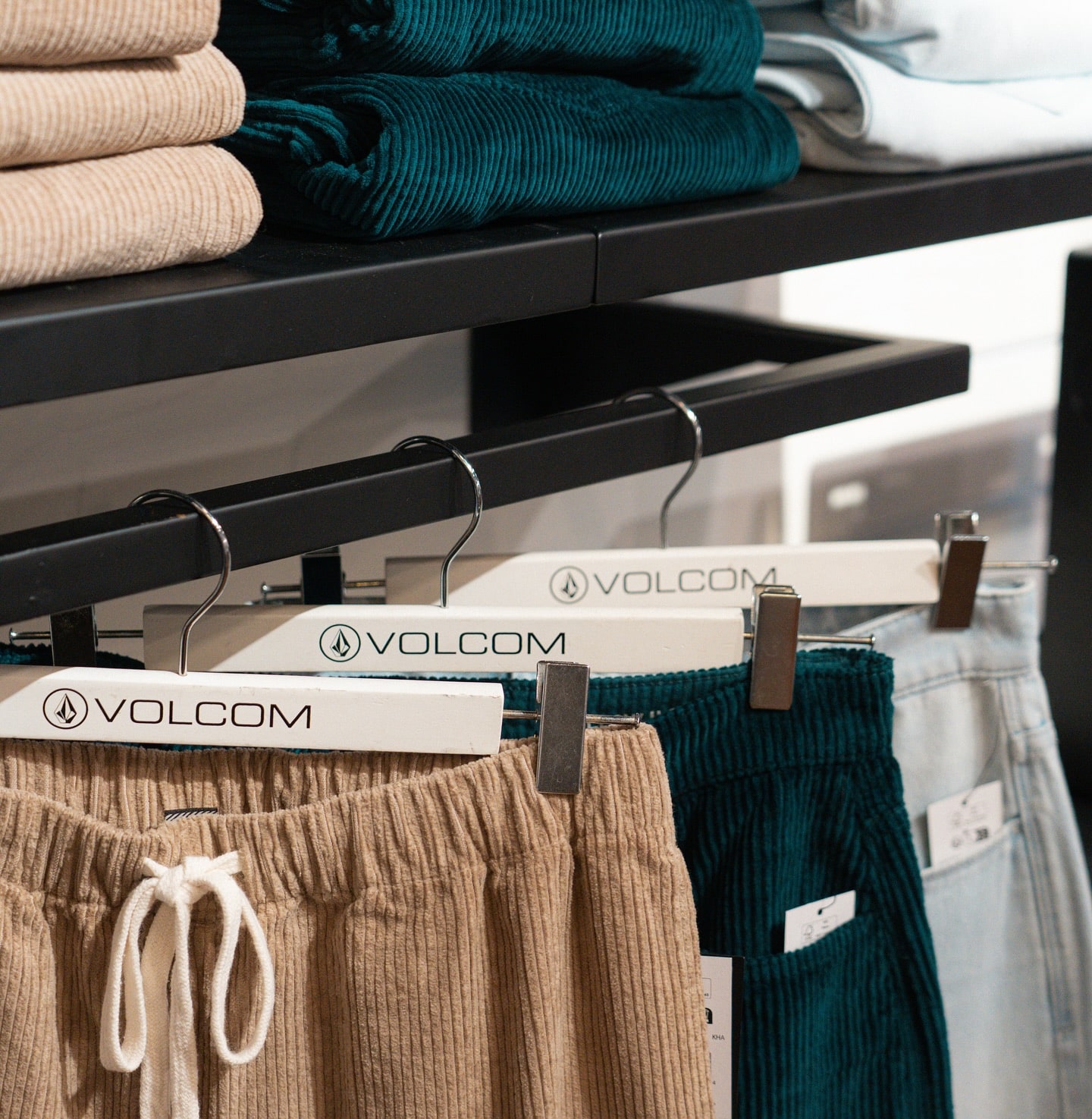 Pantalons velours côtelé beige et sarcelle suspendus sur cintres VOLCOM. Détail de la collection de vêtements sur étagère noire de magasin.