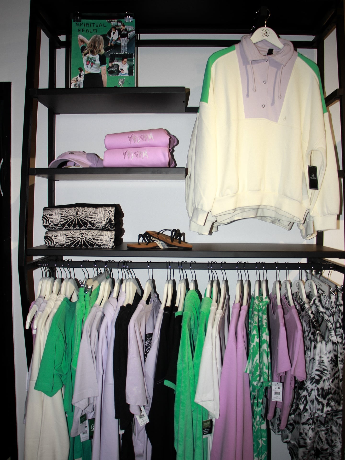 Présentoir Volcom, « SPIRITUAL REALM ». Chandail oversize crème/lilas, serviettes roses, sandales. Vêtements mode suspendus : vert, mauve, noir.