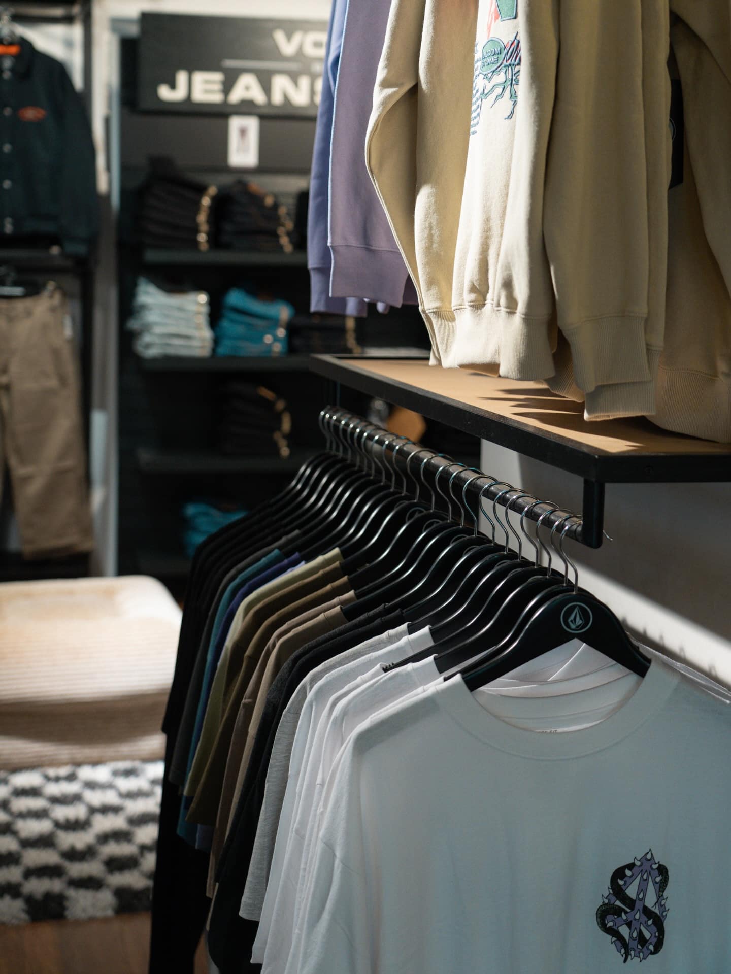 Portant de t-shirts Volcom blancs, pulls et jeans pliés dans un magasin. Hangers avec logo, panneau "VOLCOM JEANS" visible.